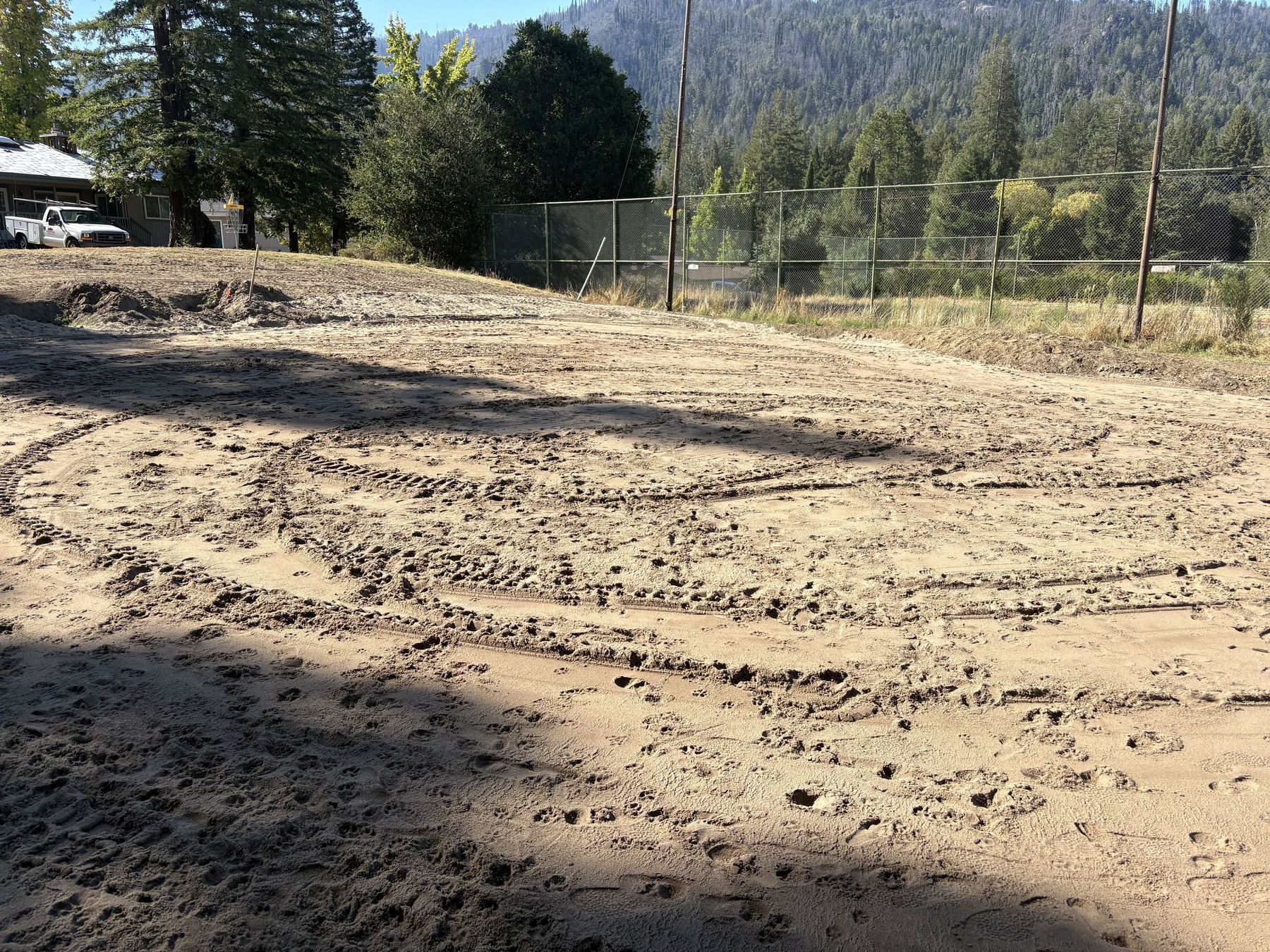 Tire tracks and footprints across the freshly graded dirt of the 12th green, with a chain-link fence, redwoods, and a white pickup beyond.