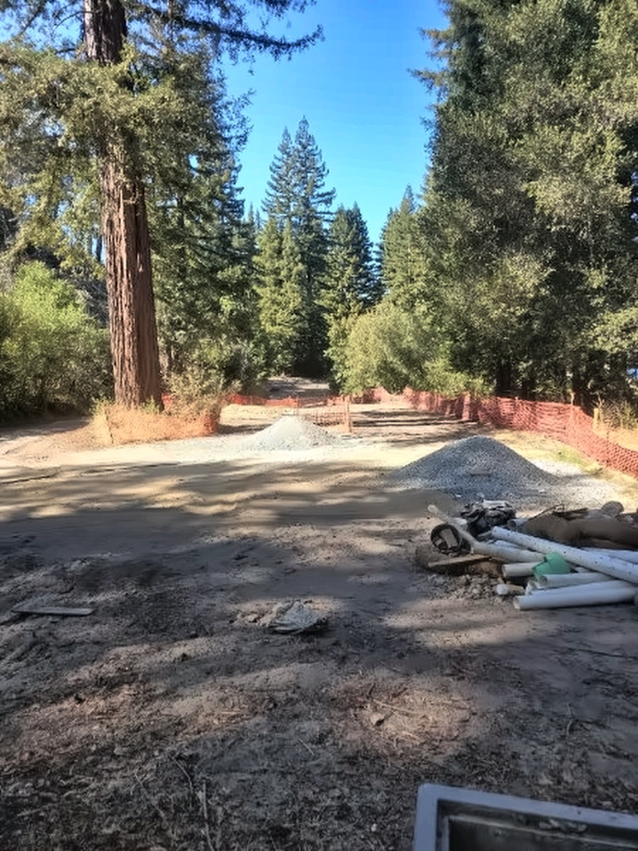 Looking out from the 18th tee box past a tall redwood, with gravel piles and supplies staged along the orange fencing