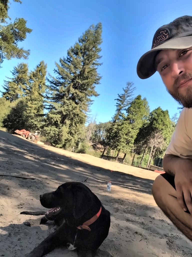 Memphis taking a selfie with his black lab Banjo on the graded 17th fairway, with a tractor and redwoods behind them