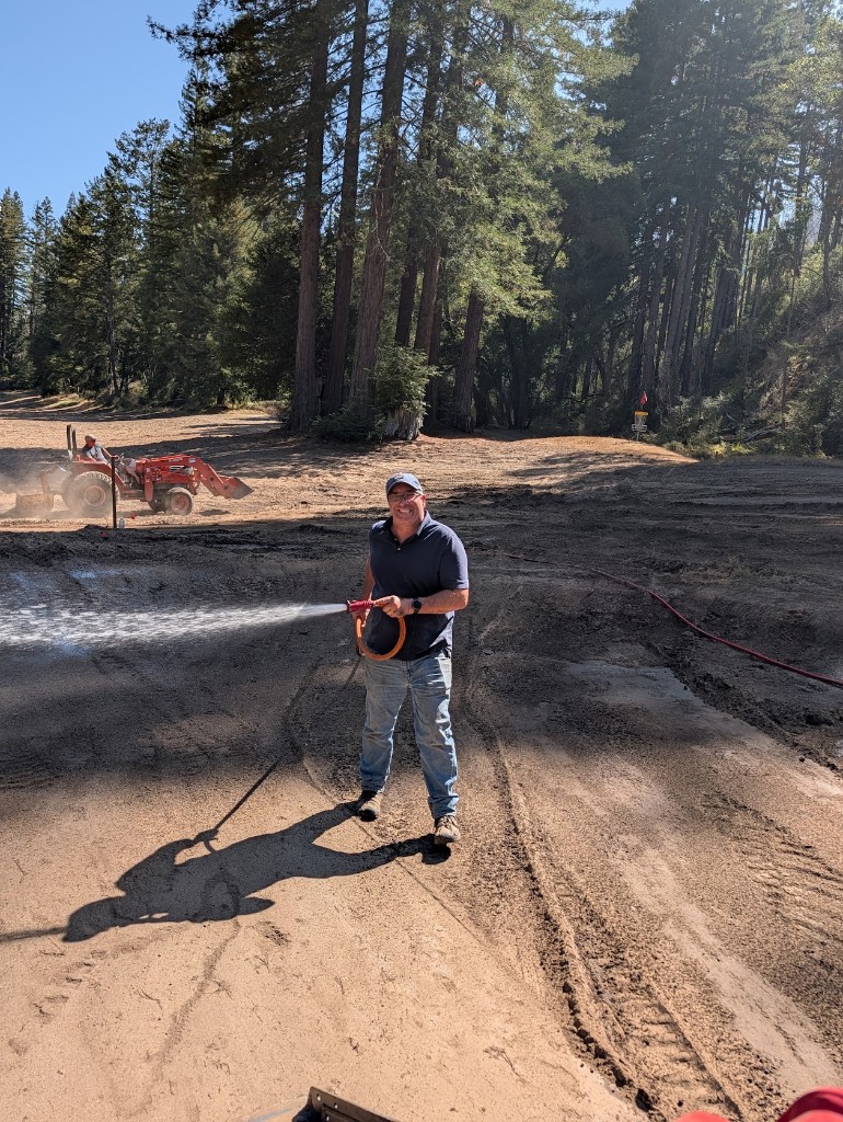 Community Crew member watering freshly graded ground on the back nine, with a tractor working behind him